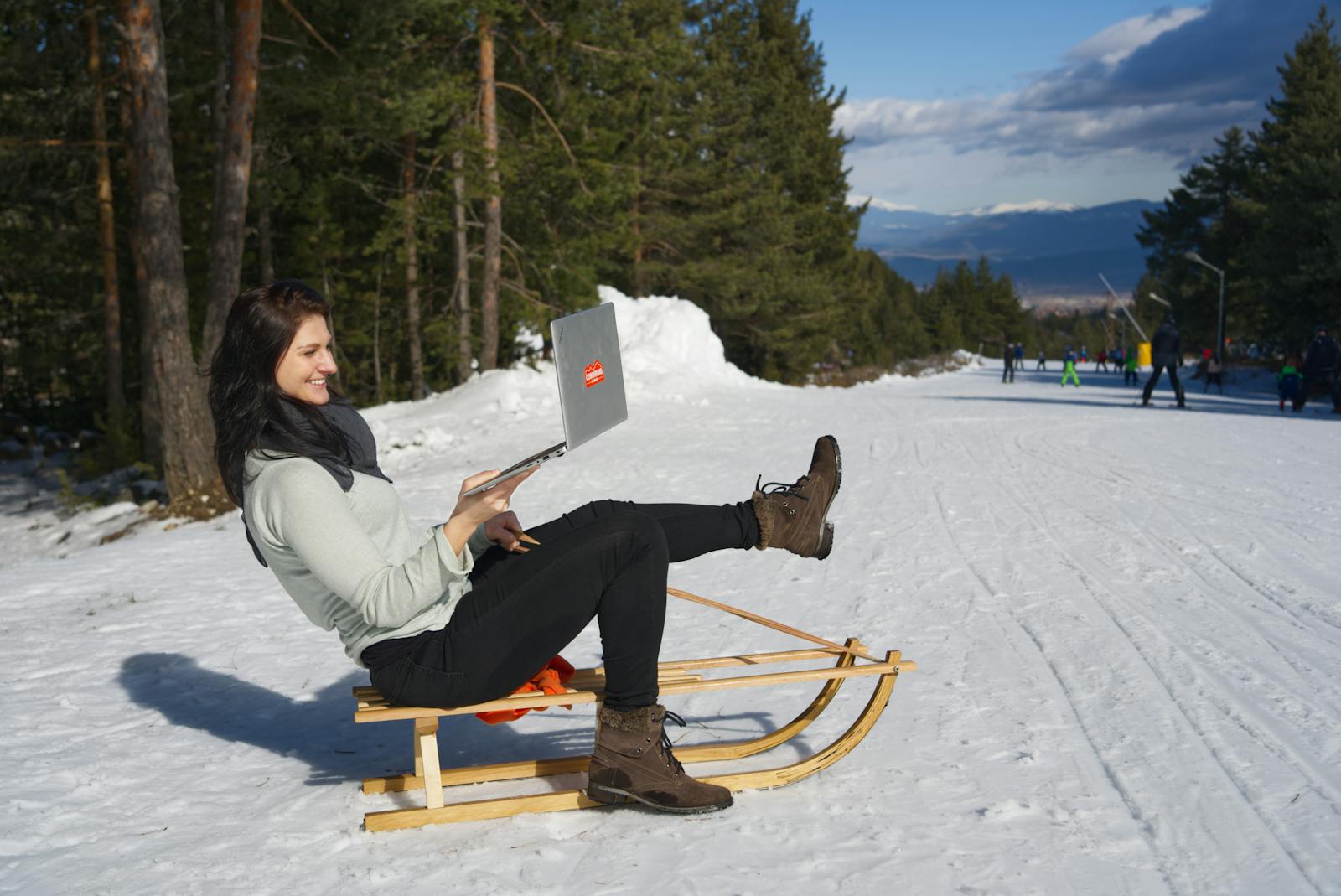 Woman using a laptop at a snowy ski resort with mountain surroundings.