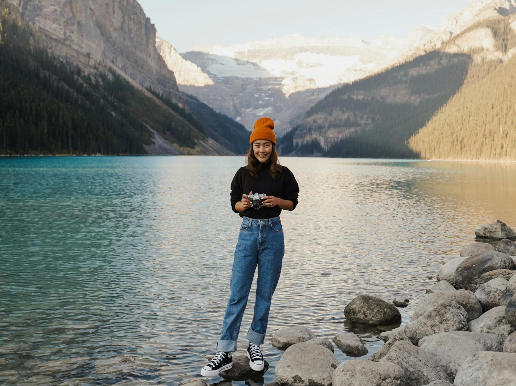 Young woman standing by Lake Louise, Canada, holding a camera, wearing casual attire.