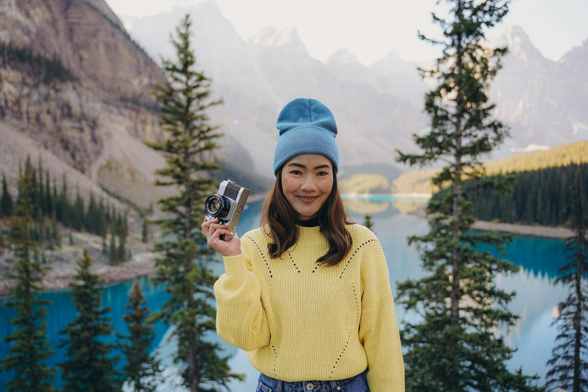 A young woman holds a camera, smiling by the vibrant blue waters of Peyto Lake, surrounded by mountains.