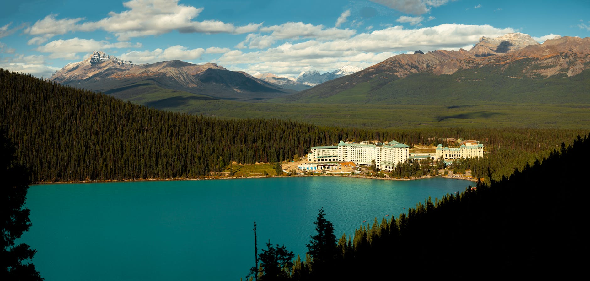 Panoramic view of a lake, forest, and hotel nestled among mountains under a clear sky.