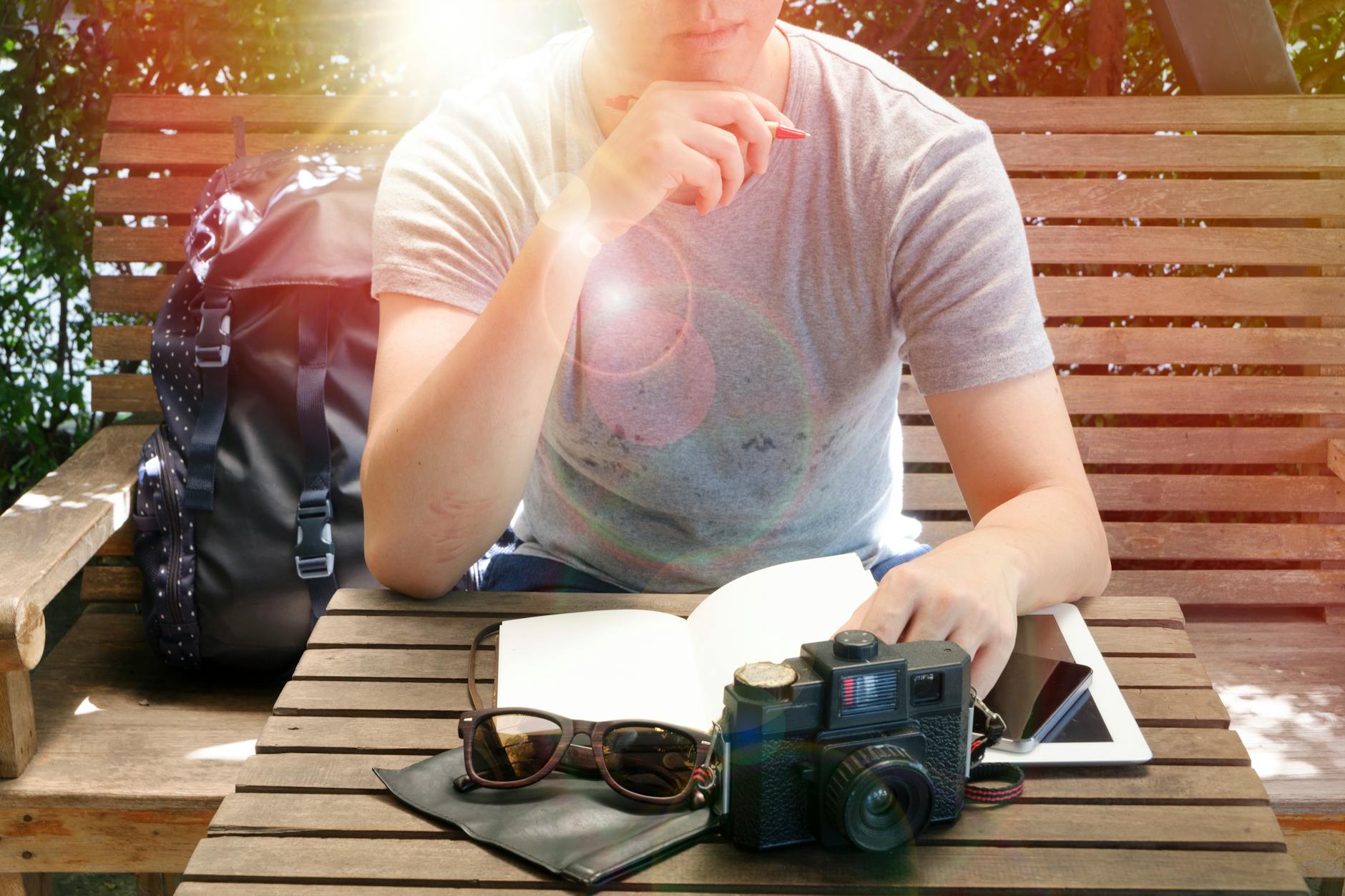 Man sitting on a wooden bench outdoors, reflecting with a camera, notebook, and sunlight surrounding him.