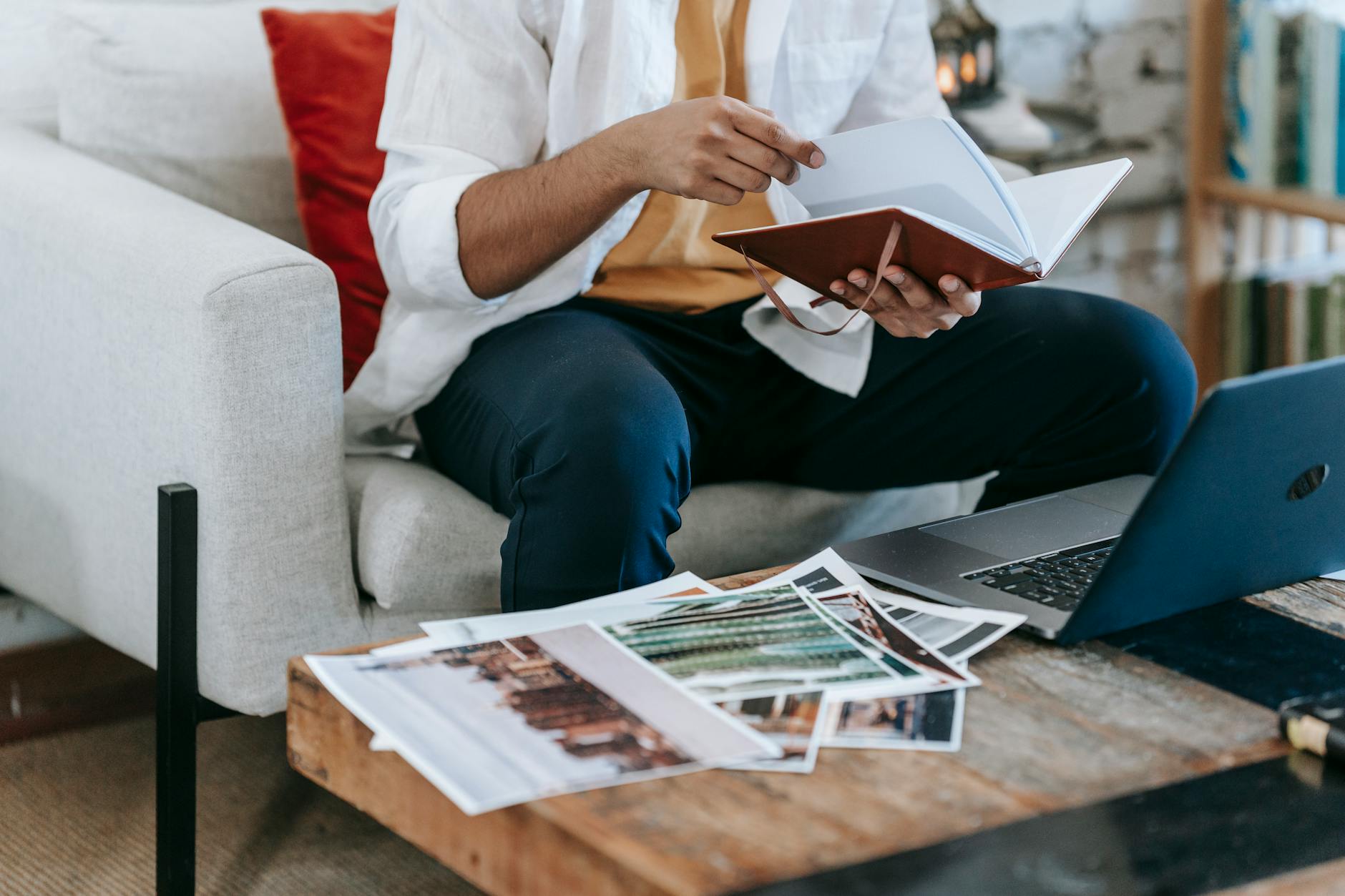 Crop anonymous male freelance designer checking notes in planner while working with laptop and printed pictures at home