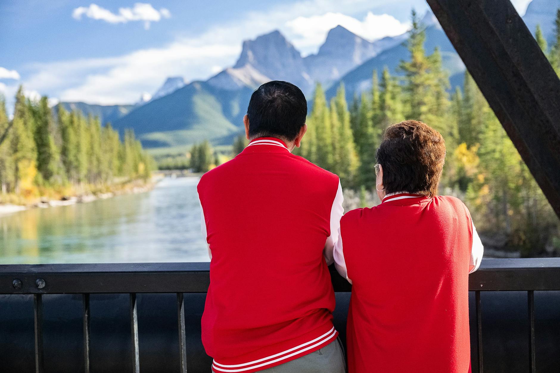 A couple in matching jackets admire mountain scenery from a bridge, surrounded by trees and a river.