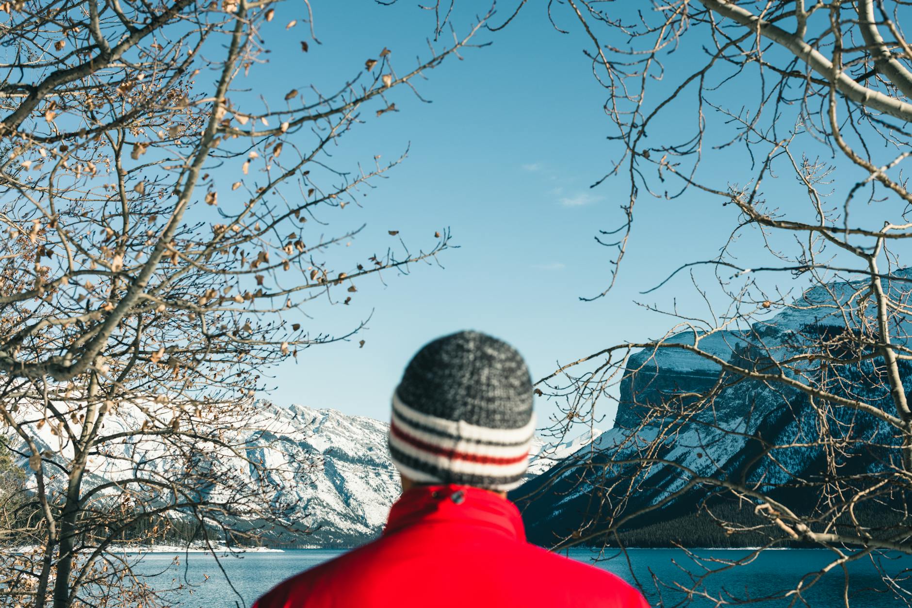 Person in red jacket and knit hat admiring snowy mountains and lake in Banff, Canada during winter.
