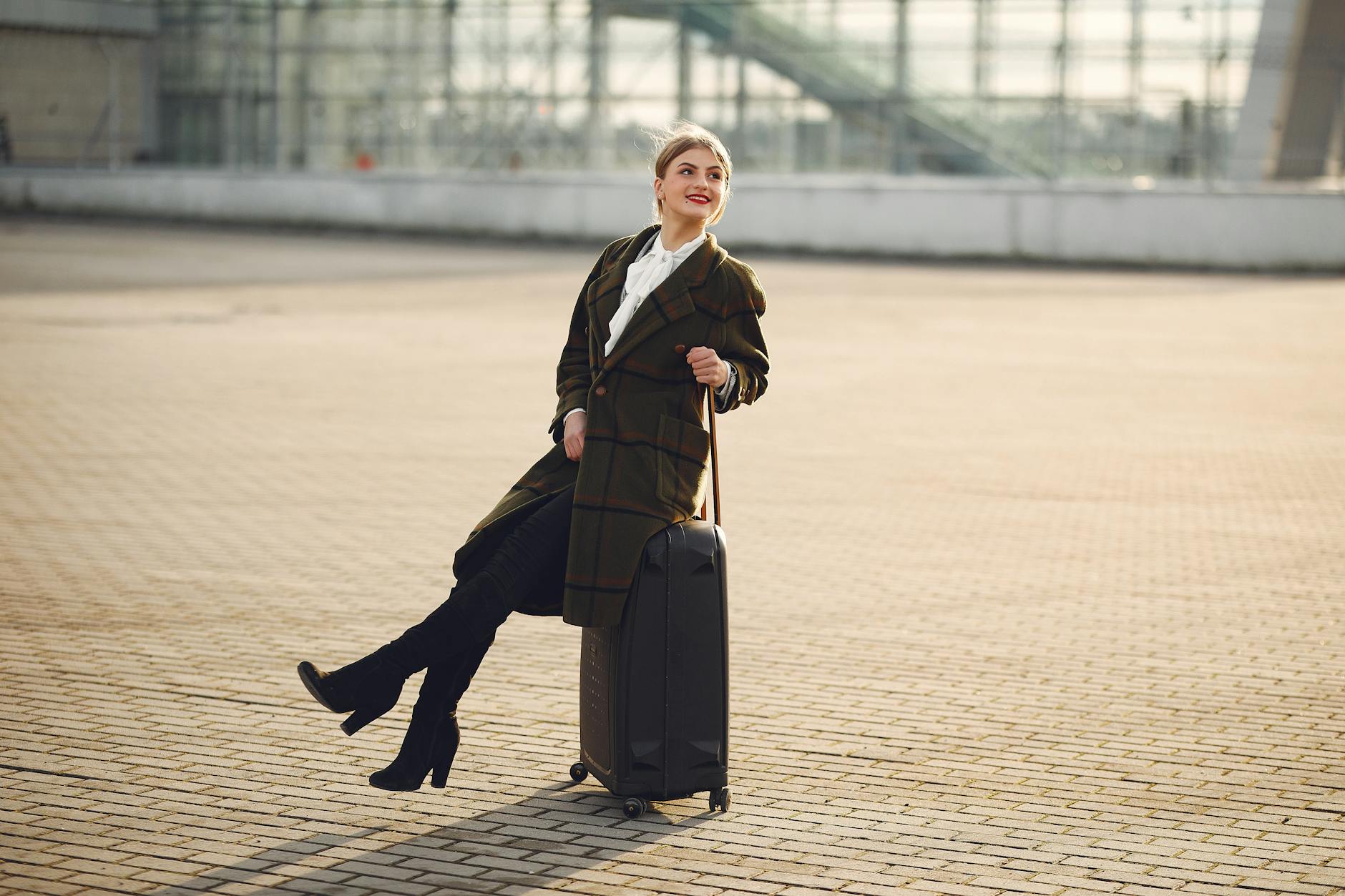 Full length of happy female traveler wearing warm trendy plaid coat and high boots sitting on top of suitcase on pavement near modern airport terminal and smiling looking away during daytime in cold season