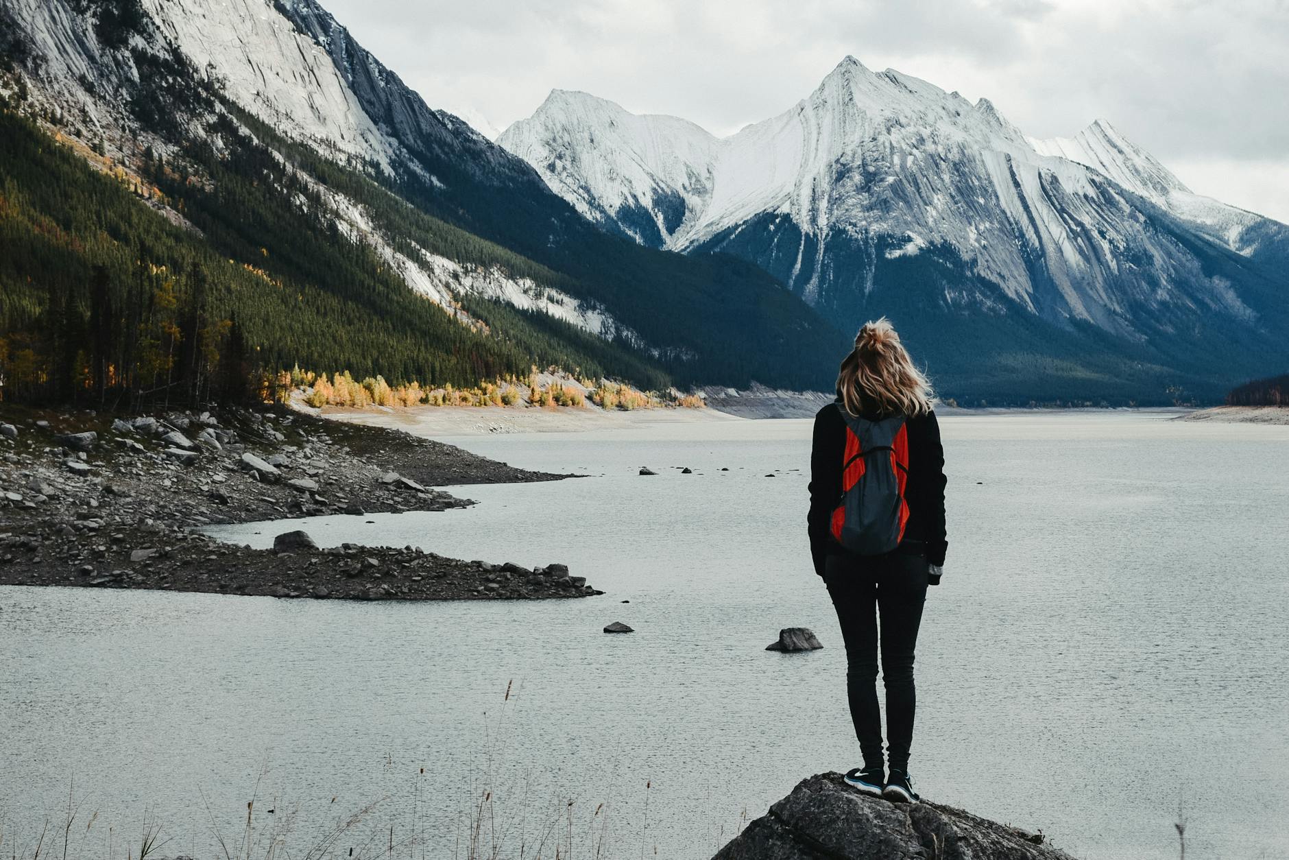 Back view of unrecognizable female traveler with backpack standing on edge of rocky shore and admiring calm river surrounded by snowy mountains in cloudy day