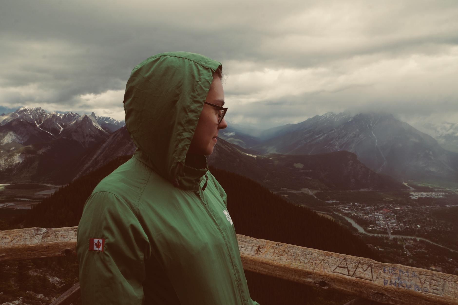 Side view of a woman wearing a green jacket with eyeglasses, gazing over a scenic mountain landscape under a cloudy sky.