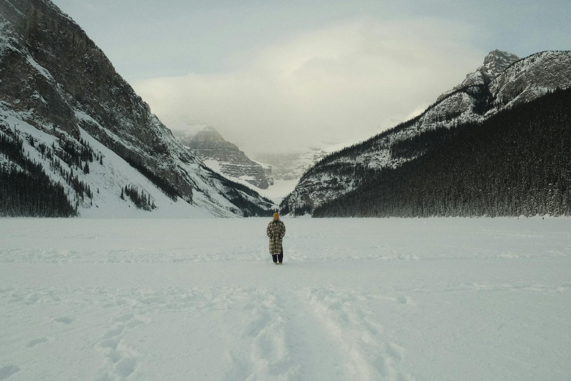 A solitary person stands in a vast snowy landscape surrounded by majestic mountains.
