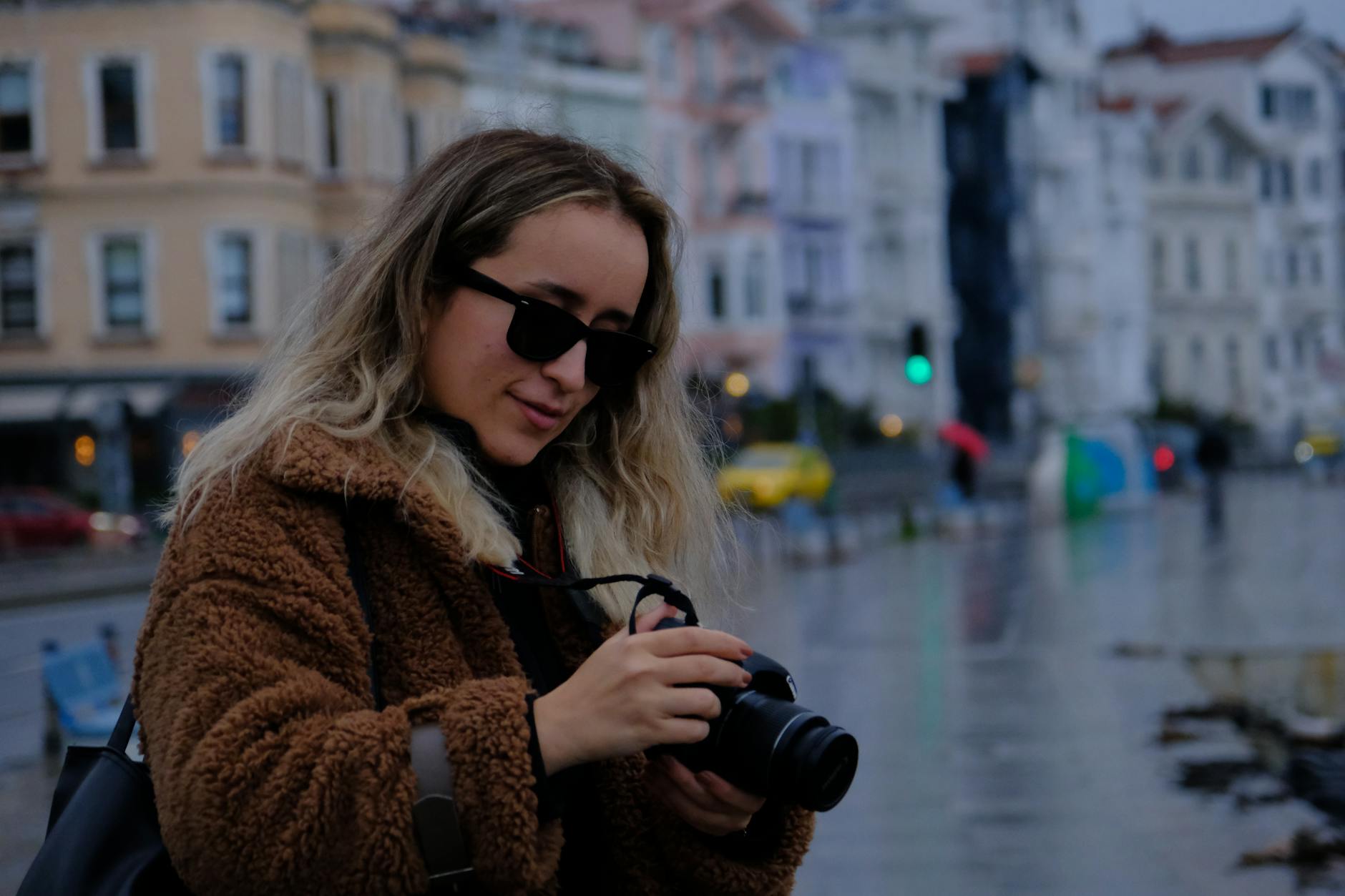 Young woman with camera in an urban street setting, wearing warm coat and sunglasses.