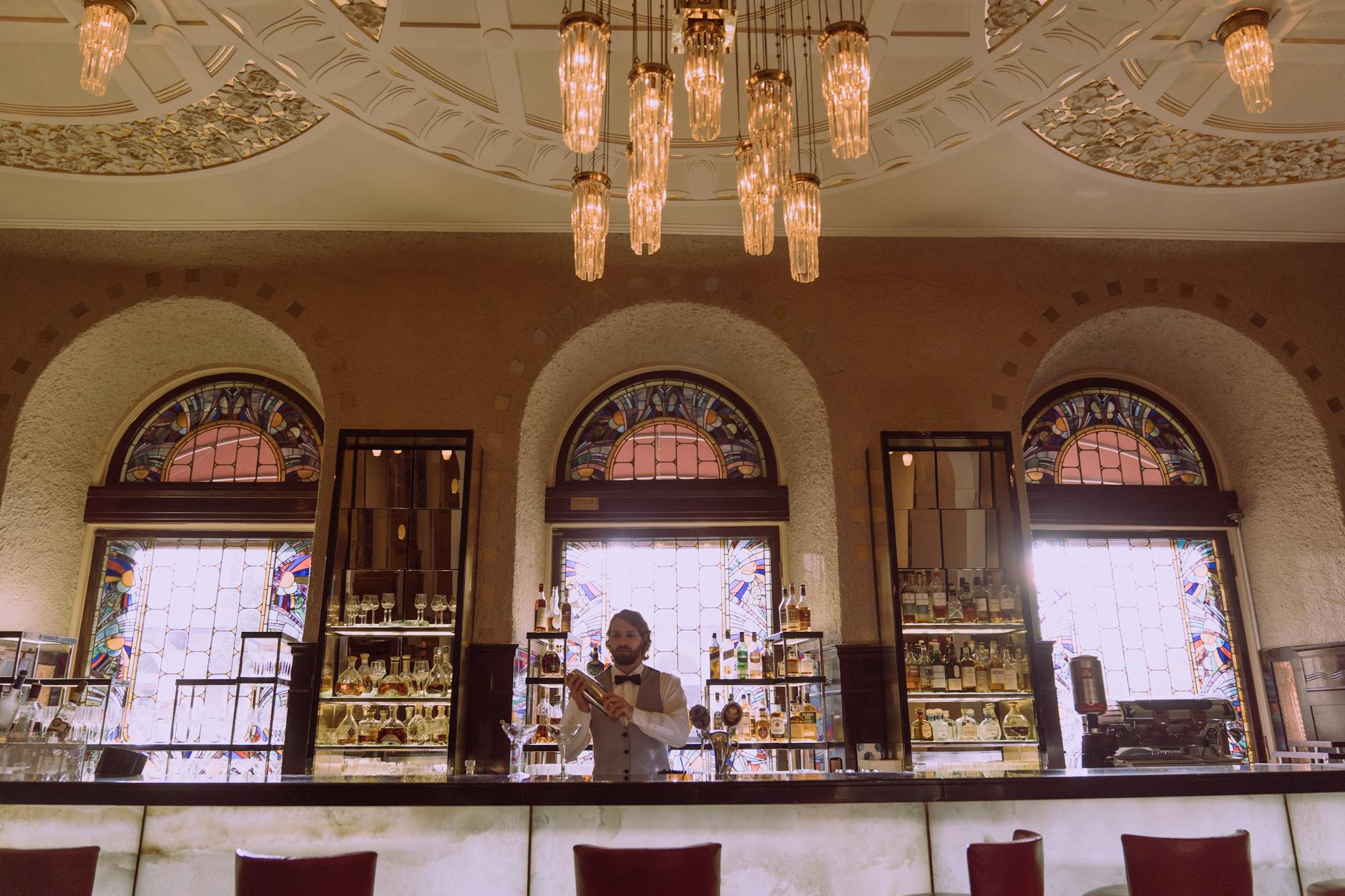 A sophisticated bar featuring a vintage chandelier and stained glass windows with a bartender serving drinks.