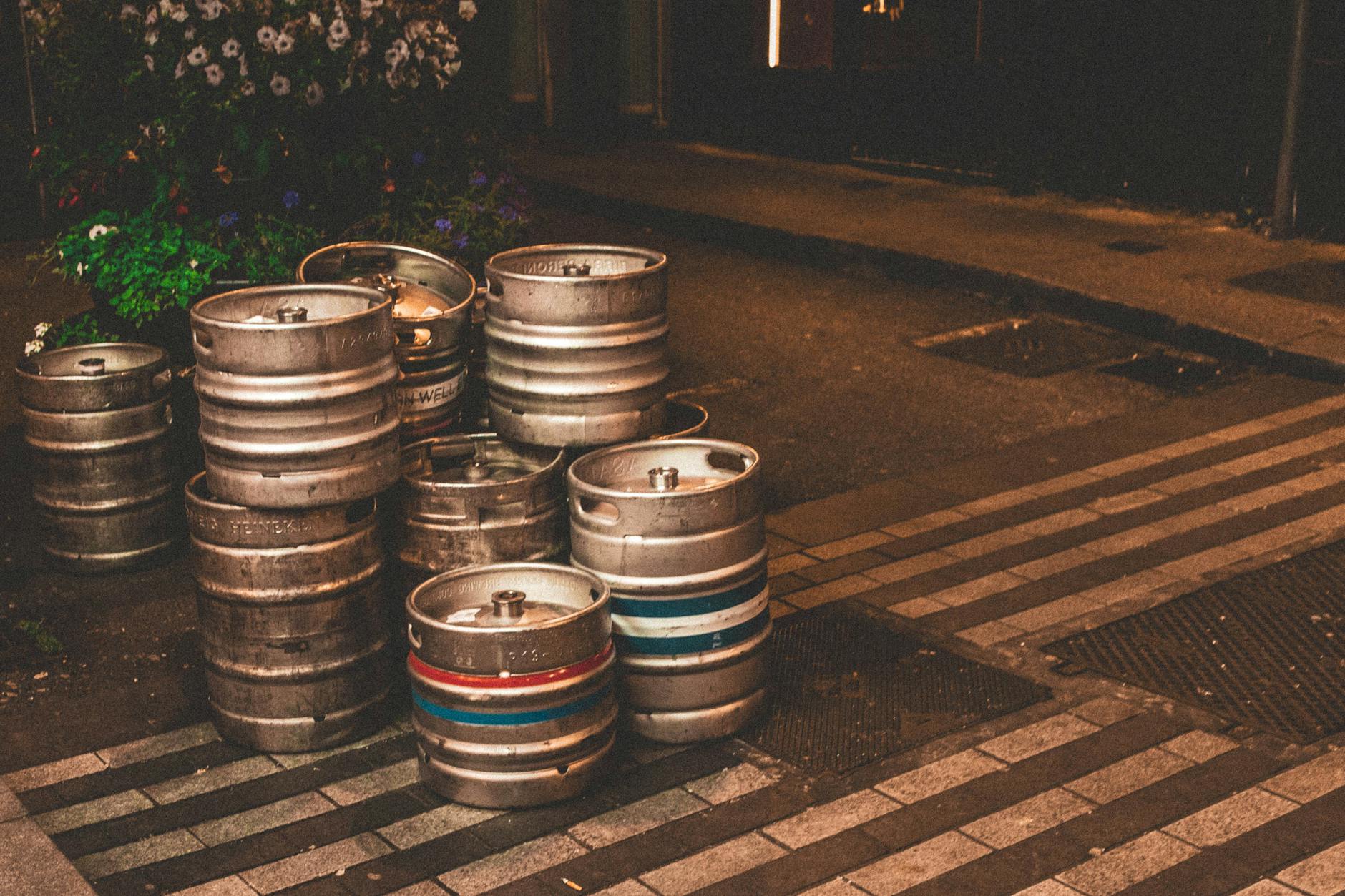 Rustic street scene with stacked metal kegs outside a pub entrance on a cobbled sidewalk.