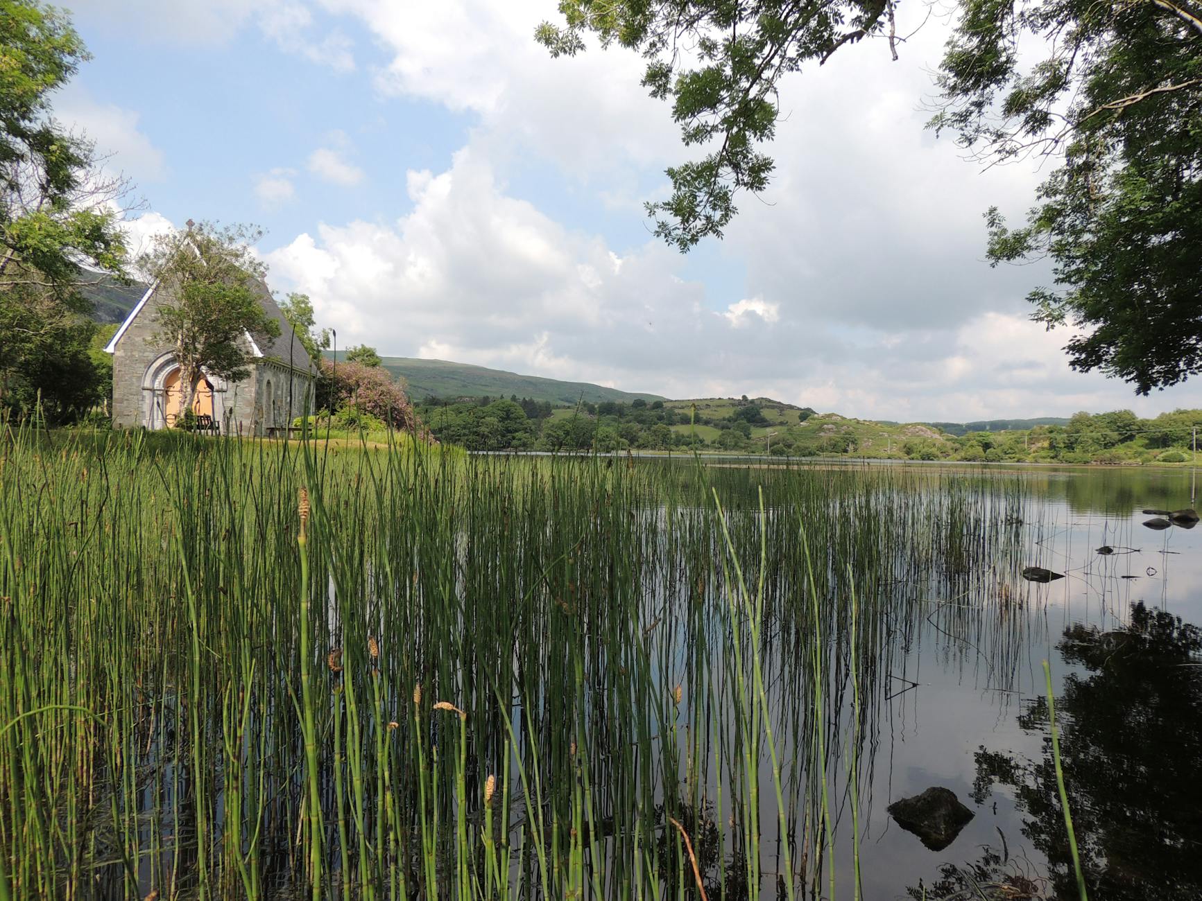 A picturesque stone chapel by a serene lake surrounded by lush greenery and hills in Ireland.