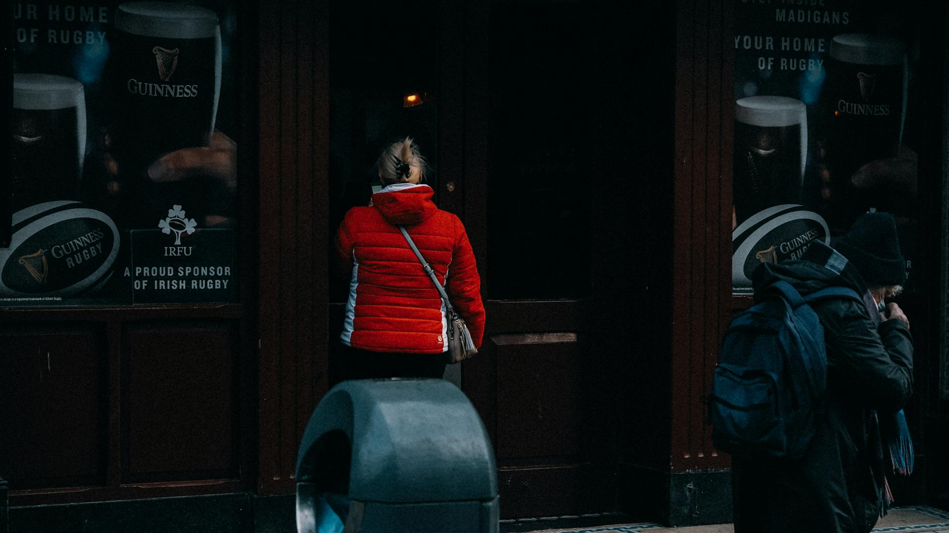 A woman in a red jacket stands at a pub door, viewed from behind on a city street.