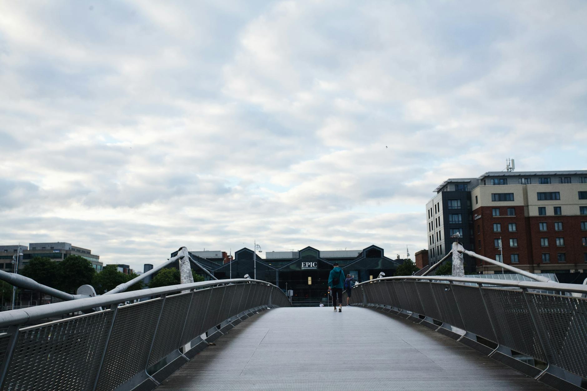 Person walking on a bridge with buildings and cloudy sky in background, urban scene.
