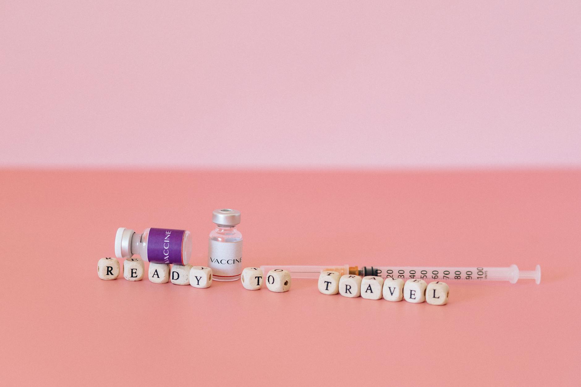 Close-up of vaccine vials, syringe, and the words Ready to Travel on a pink background.