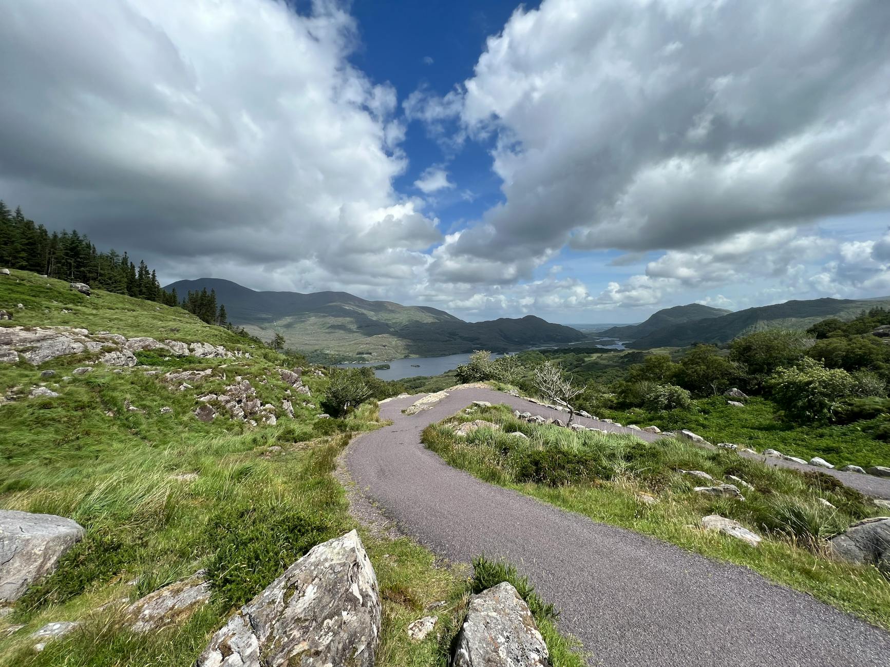 Winding road through lush green valley with mountains under cloudy sky.