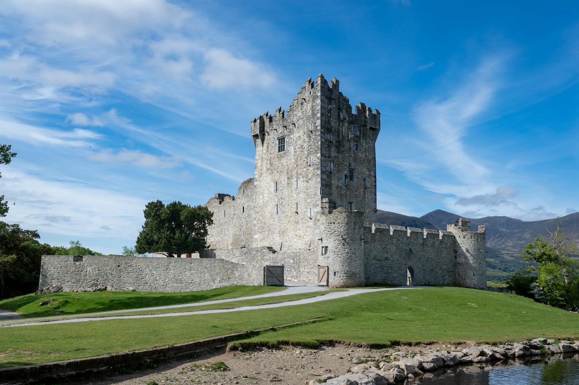 A scenic view of Ross Castle, Killarney, Ireland, under clear blue skies with green landscape.