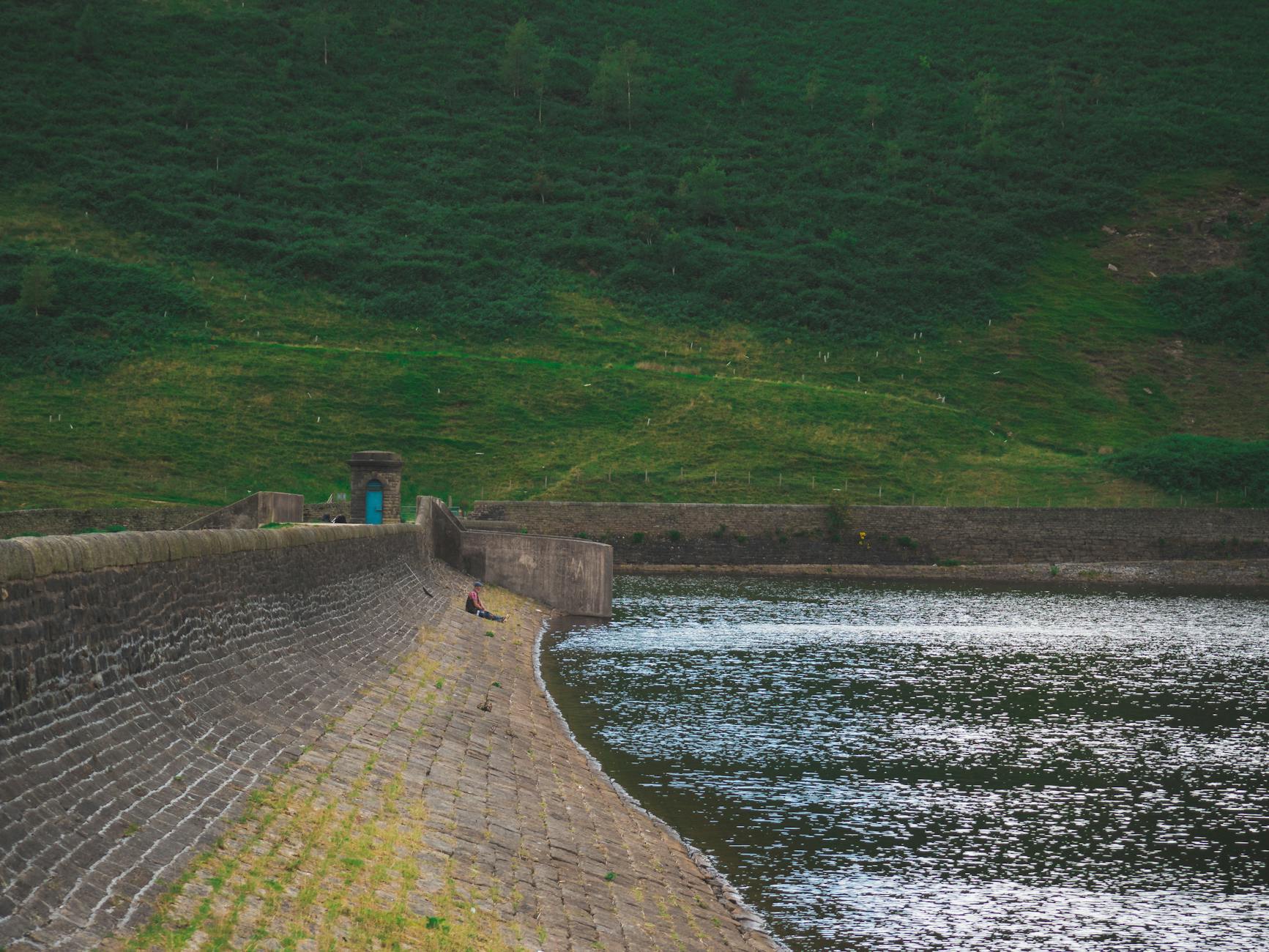 Person sitting by a reservoir dam surrounded by green hills