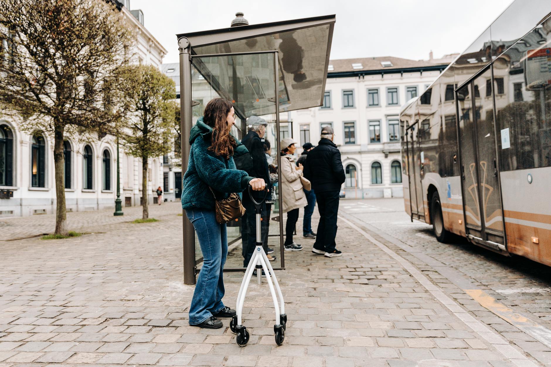 People waiting at a city bus stop, including a person using a rollator