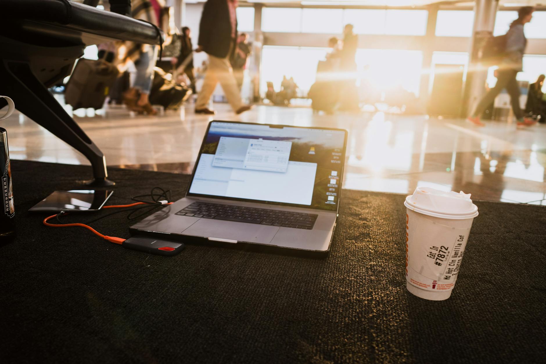 Laptop and coffee cup on an airport floor with passengers nearby