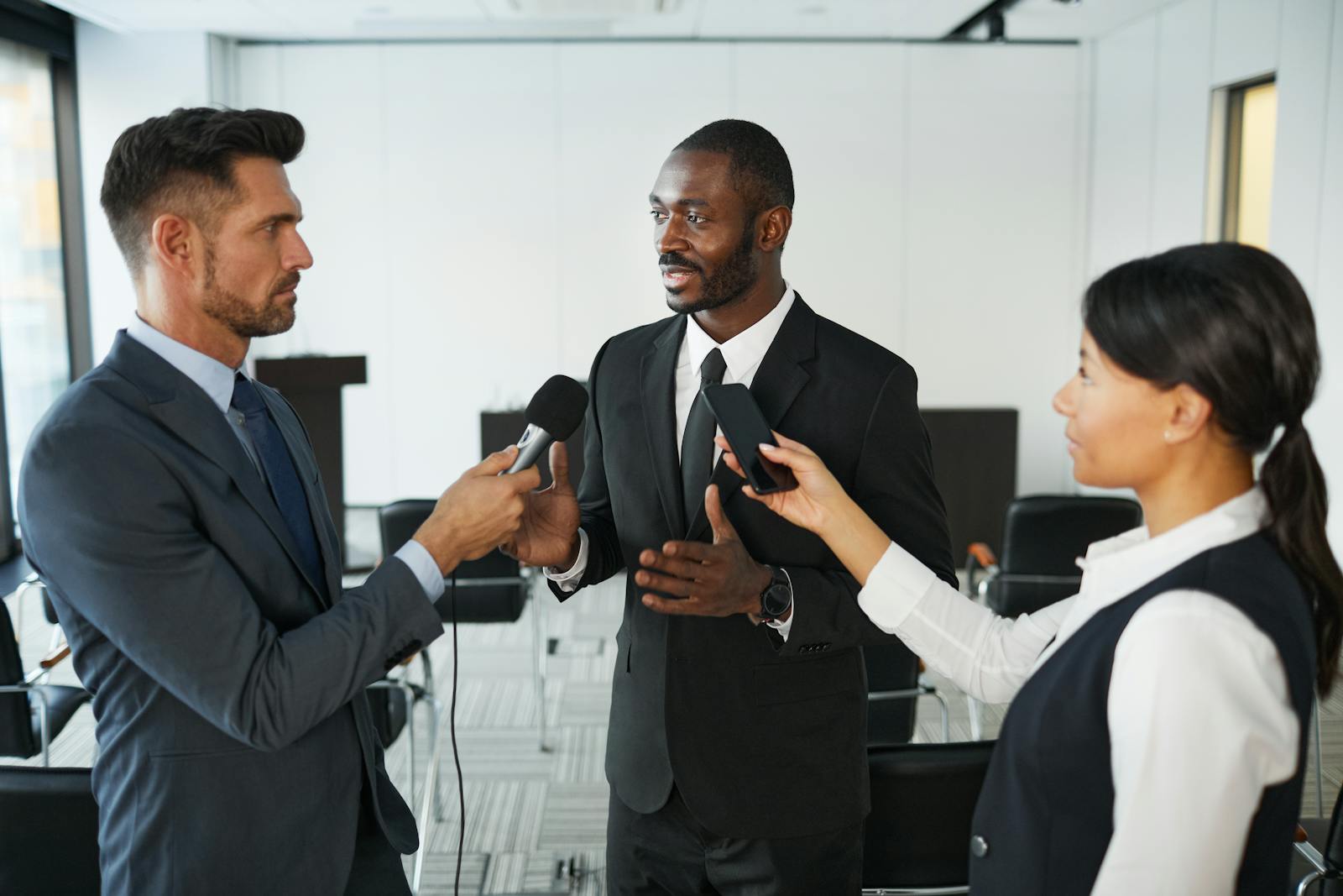 Businessman in formal attire being interviewed by journalists with microphones in a conference room.