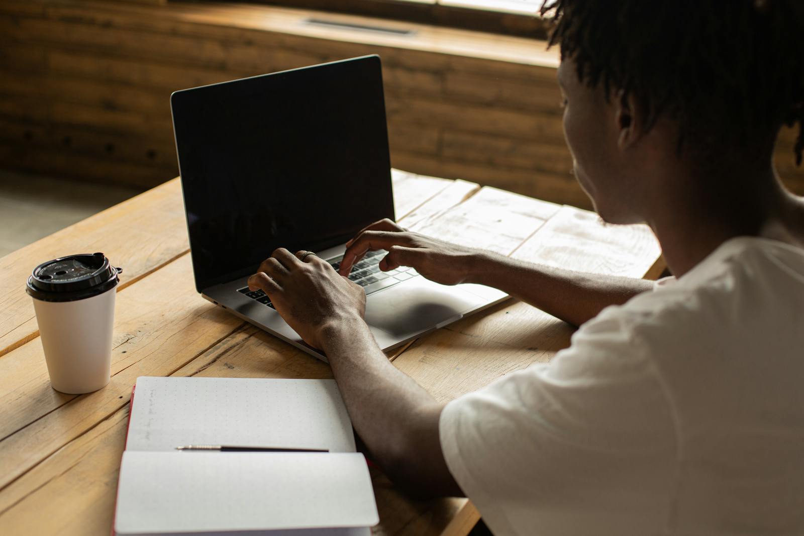 Young man working on a laptop in a cozy cafe with coffee and notebook on table.