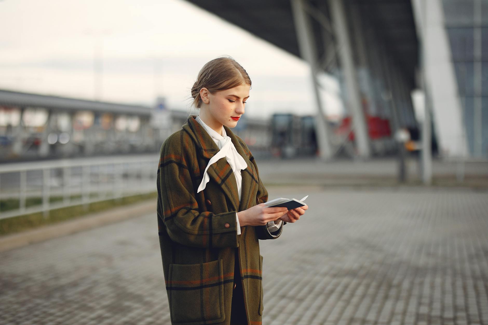 Attentive female passenger wearing trendy plaid coat and white blouse checking passport and ticket standing on pavement near modern building of airport out
