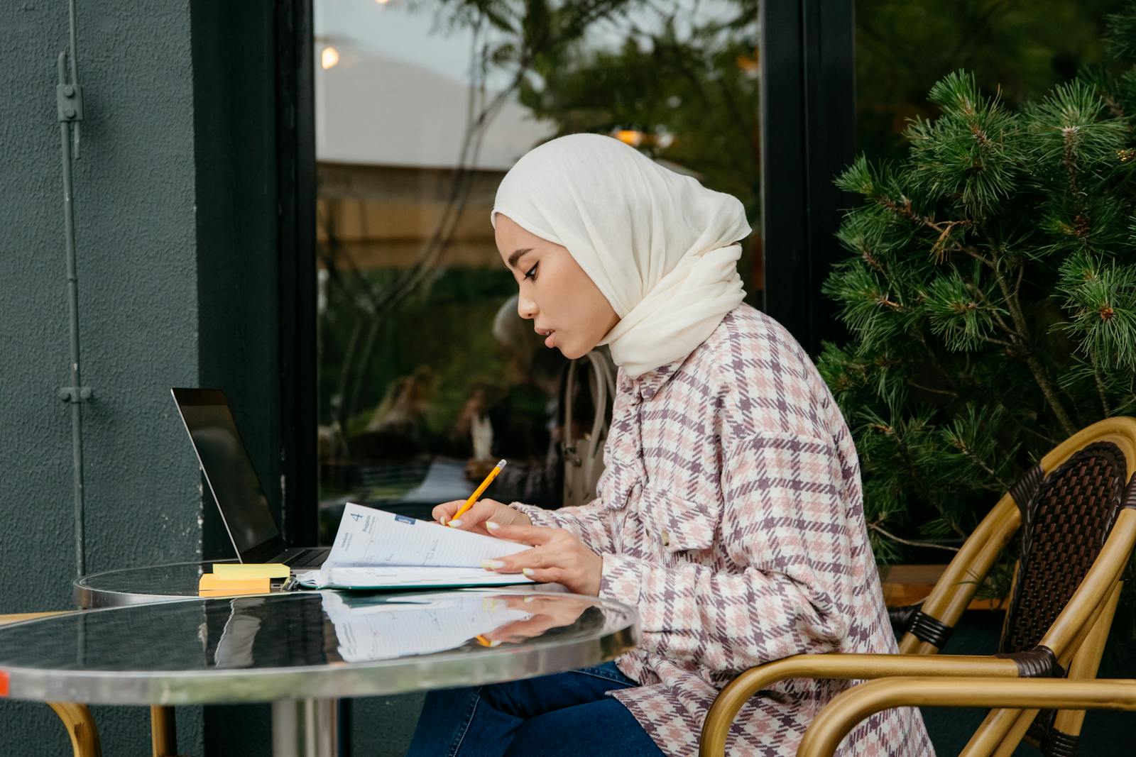 Young Muslim woman wearing hijab studying with laptop and notebook at an outdoor café table. Productive and focused.