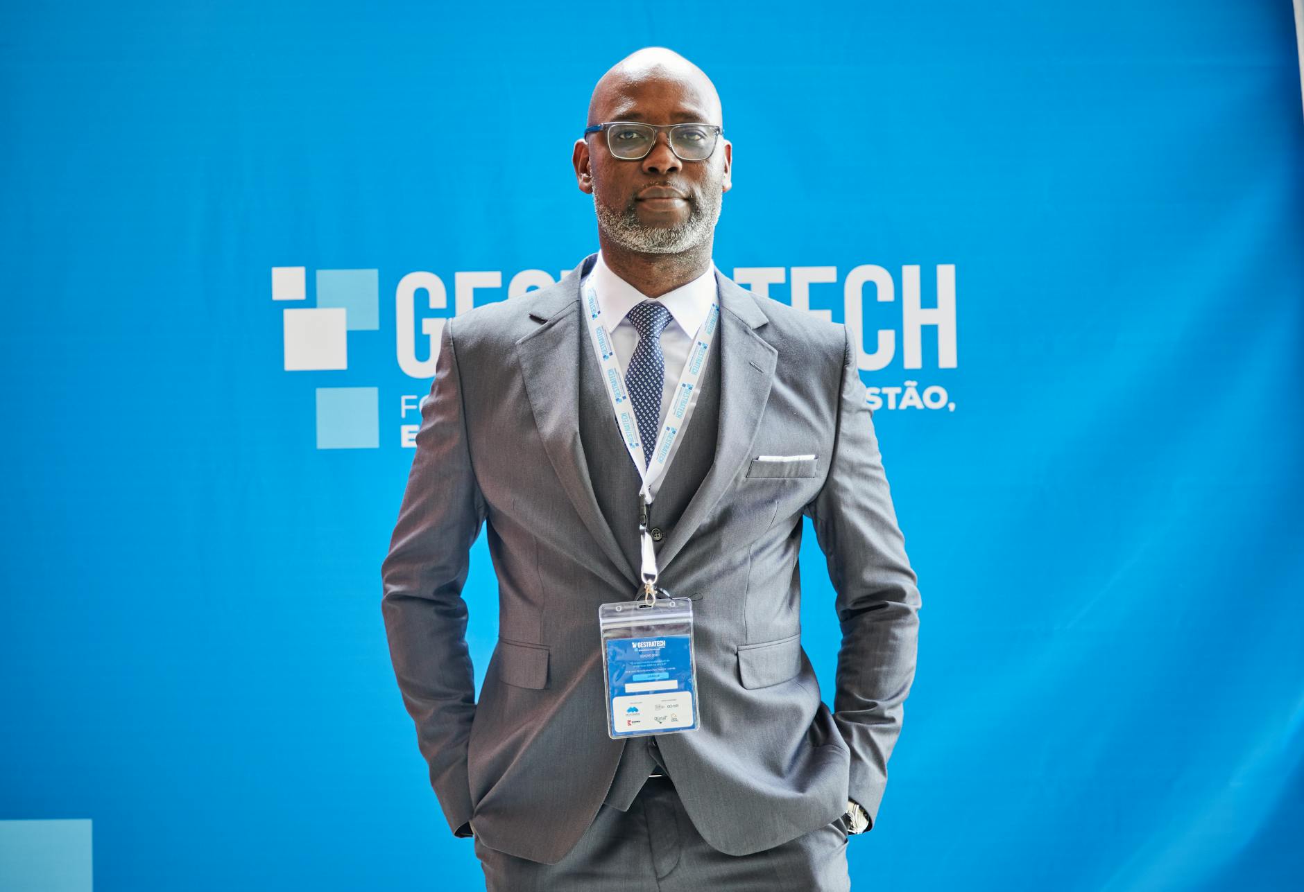 Confident African man in a grey suit at a trade fair with blue photo wall backdrop.