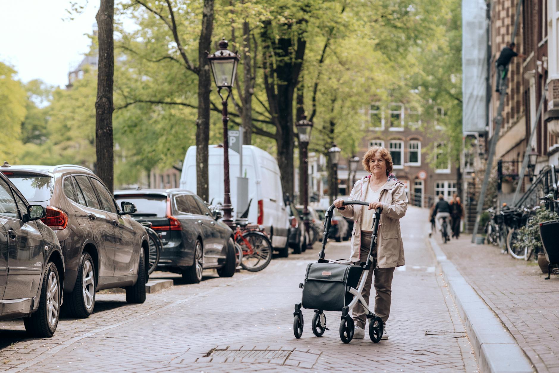 Elderly woman strolling along cobblestone street in Amsterdam with a walker, surrounded by parked cars.