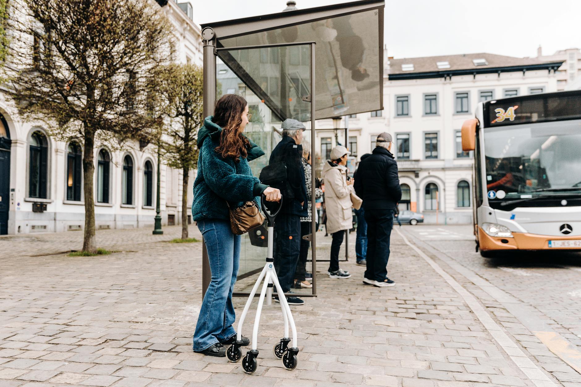 A diverse group waits at a city bus stop, including a young woman with a rollator.