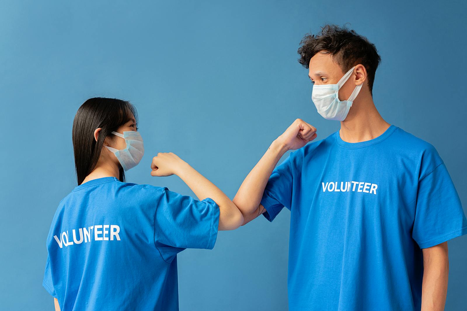 Two volunteers wearing blue shirts and face masks elbow bump for safety.